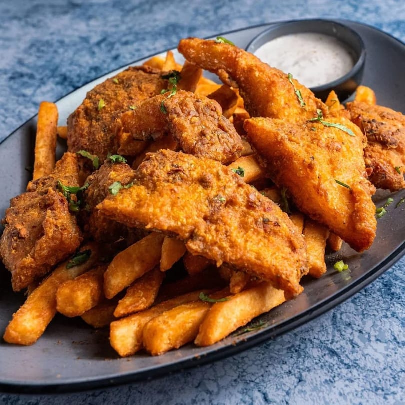 Crispy golden-brown fish pakora and thick-cut fries with a side of white dipping sauce on a dark plate.