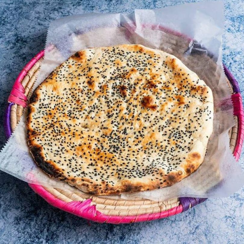 A round, golden-brown flatbread topped with black sesame seeds, served in a colorful woven basket.