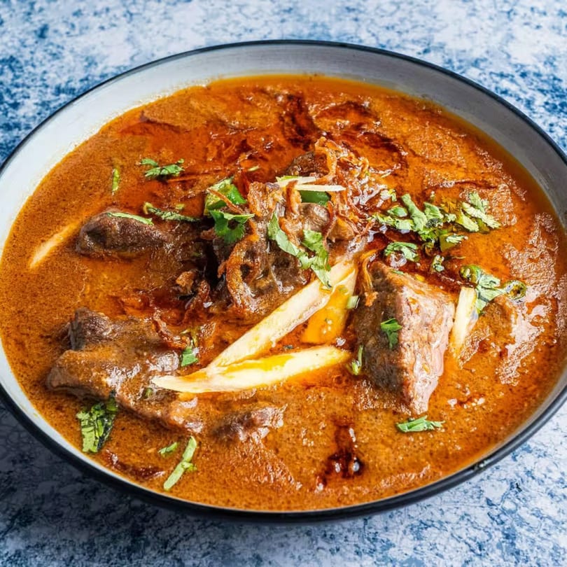 Close-up of a rich, orange-brown Pakistani curry with tender meat, garnished with cilantro and ginger.