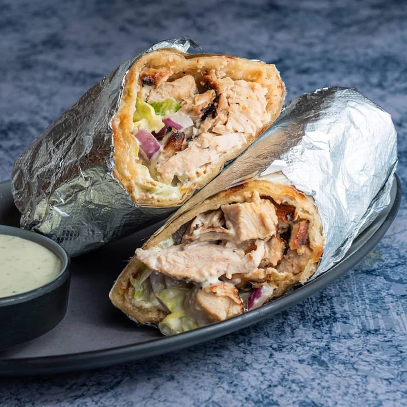 Two foil-wrapped chicken shawarmas, cut in half, revealing meat, lettuce, and red onion, with a side of sauce.