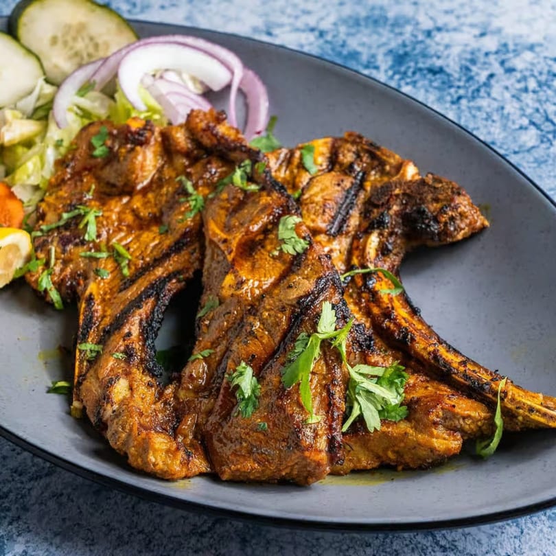 Grilled lamb chops with fresh parsley, sliced onions, cucumbers, and lemon on a dark plate.