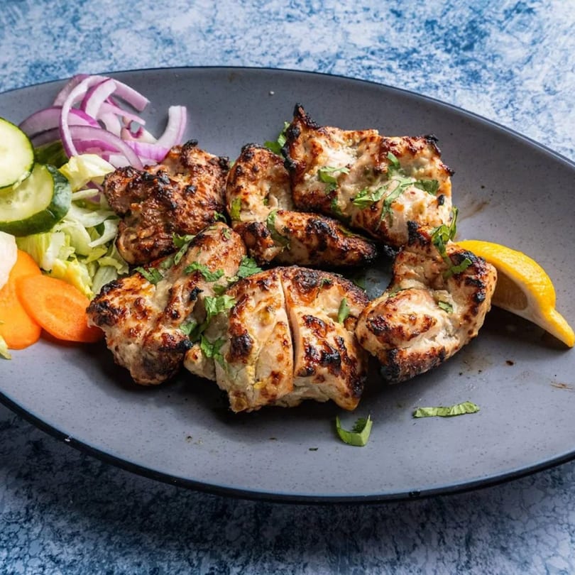 Grilled chicken tikka pieces with charred edges, garnished with cilantro, served with salad and a lemon wedge.