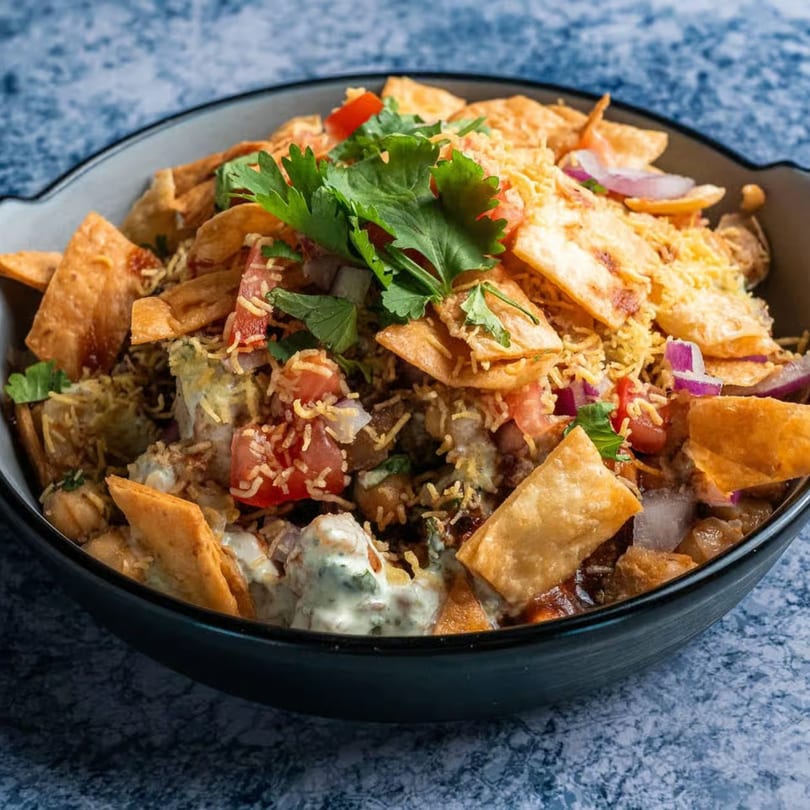 A close-up of a bowl of Pakistani chaat with crispy flatbread, tomatoes, onions, and cilantro.